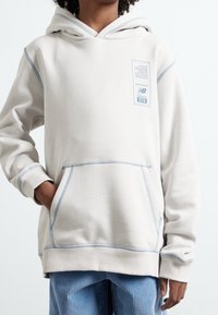 Kremowa bluza z kapturem z kontrastowym jasnoniebieskim przeszyciem, kieszenią typu kangur i małą naszywką z logo na klatce piersiowej. Miękki, teksturowany materiał bawełniany.