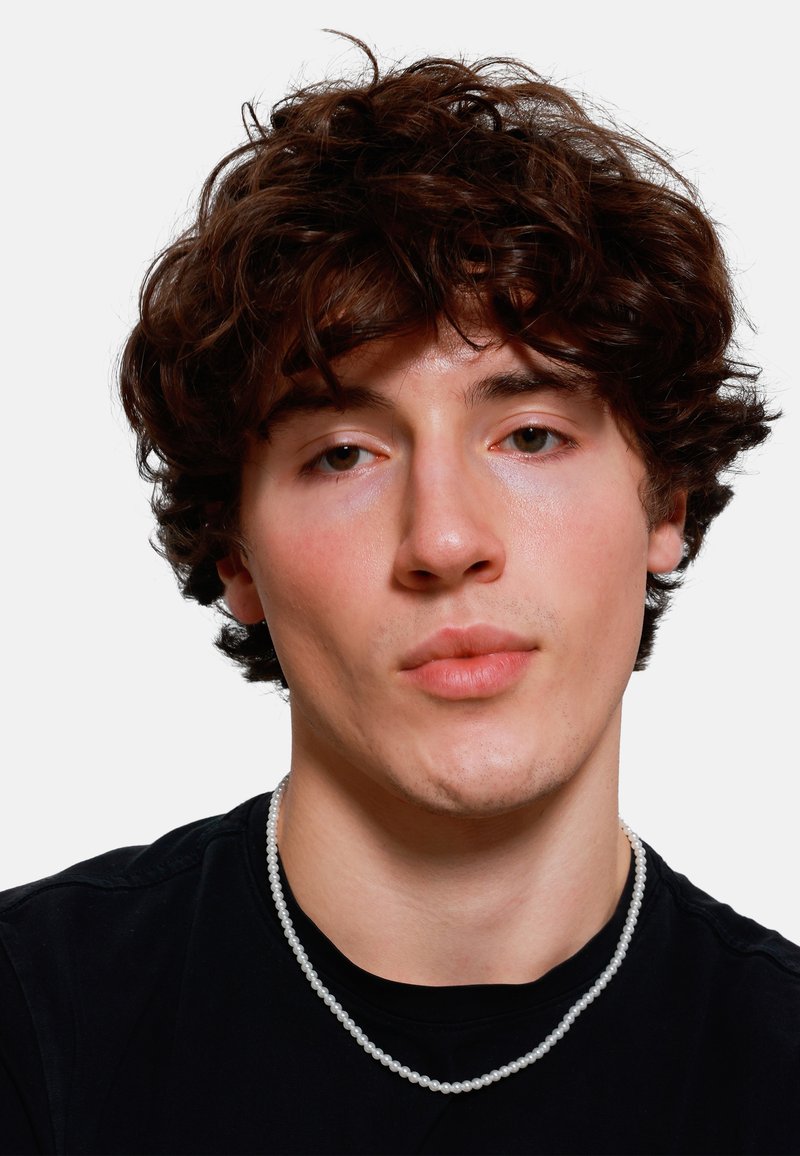 Portrait of a person wearing a black T-shirt and a white pearl necklace. Features curly brown hair and smooth complexion. Neutral background.