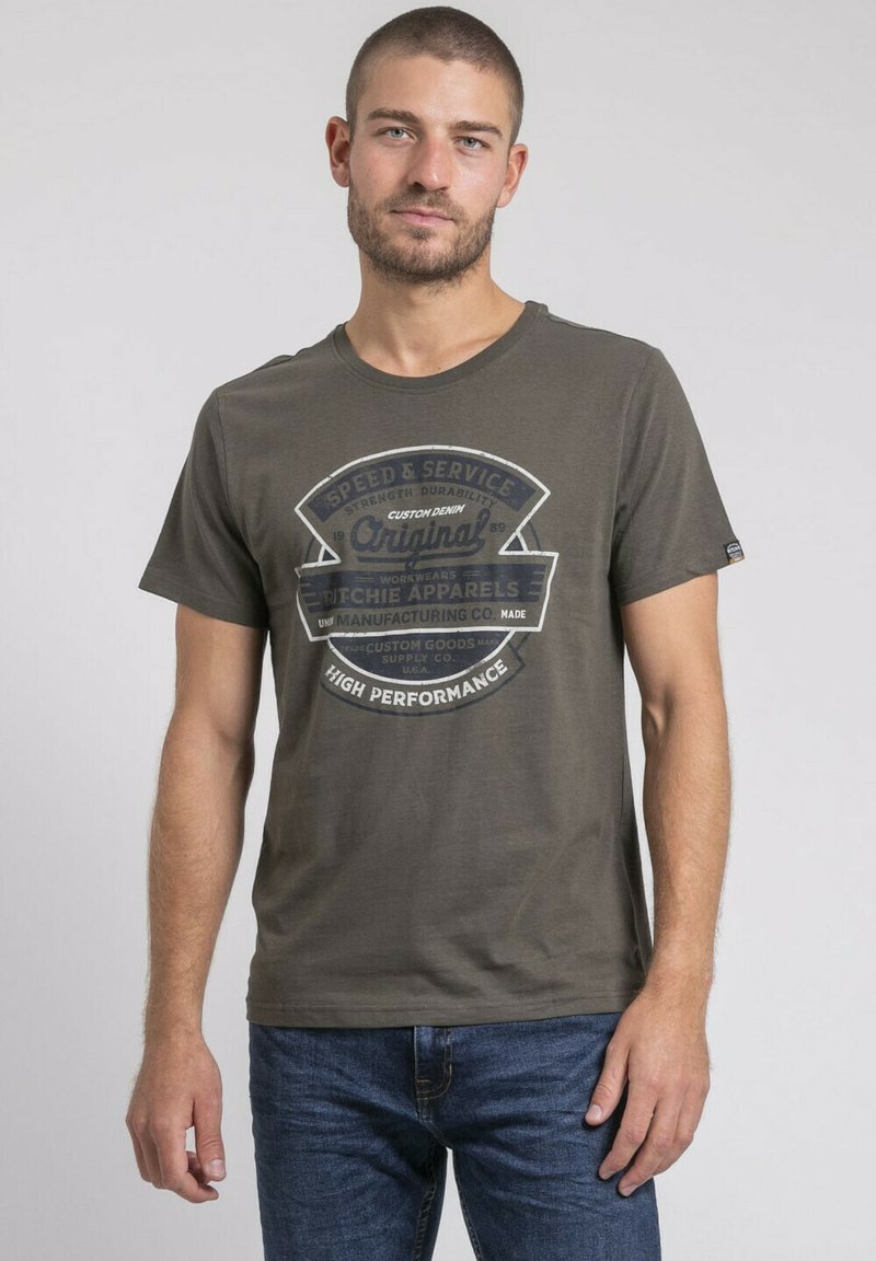 T-shirt en coton vert olive avec un grand imprimé graphique incluant du texte et un motif circulaire en bleu foncé et blanc. Encolure classique et manches courtes.