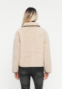 Beige fuzzy jacket with a collared neck, featuring a textured fabric and black trim along the collar and hem. Worn with dark pants.