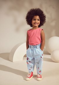 Raudonas rankovių neturintis viršus, šviesiai mėlynos dryžuotos kelnės su vaisių siuvinėjimu ir raudoni sportbačiai. Vaikas stovi kišenėse rankomis, besišypsantis.