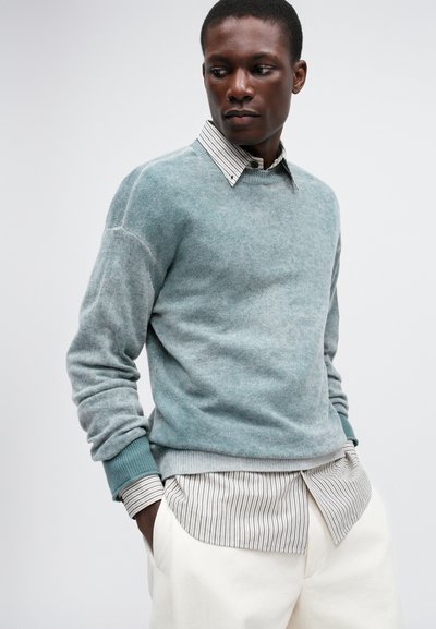 Hele teal'värvi kudumisega sviiter, mille varrukad on ribatud, on kihitatud triibulise särgi peale. Valged püksid, millel on mugav istuvus. Lihtne, tekstuuriga disain.