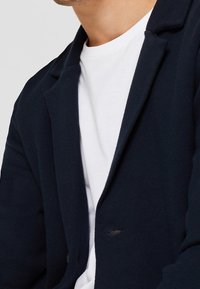 Mörkblå stickad blazer med krage, enknapplockning och textilstruktur. Bärs över en vit T-shirt som visar på lager-styling.