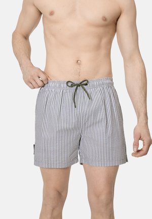 Hombre con pantalones cortos de baño de rayas verticales grises y blancas con cordón verde, ajustándose la cintura con la mano izquierda.