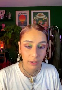 Young person with slicked-back hair, green and purple eye makeup, large earrings, layered necklaces, white shirt, colorful posters, plants, and clothing rack behind.