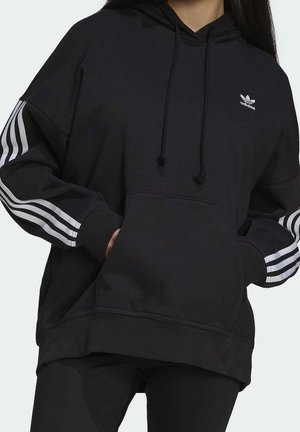 Person trägt schwarzen Adidas-Hoodie mit weißen drei Streifen an den Ärmeln und Adidas-Logo auf der linken Brust, Hände in der vorderen Tasche.
