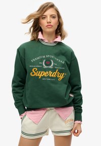 Sudadera verde con logotipo y texto, superpuesta a una camisa de cuello rosa. Pantalones cortos de marfil con rayas verdes y detalle del logotipo en el dobladillo.