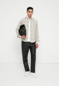 Hombre vestido con una camisa de cuadros beige sobre un suéter blanco con cremallera, pantalones de piel negros, zapatillas blancas, sosteniendo un bolso de piel negro frente a un fondo blanco.