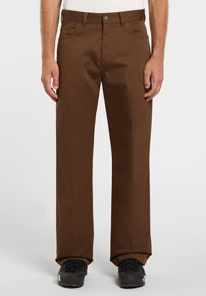 Pantalones marrones de mezcla de algodón con piernas rectas, bolsillos delanteros visibles y cierre con botón metálico. Combinados con zapatos negros para crear contraste.