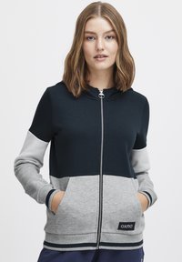 Sweatshirt à capuche zippé avec un mélange de couleurs bleu marine et gris, des poignets côtelés et une poche avant avec un logo d'accent. Texture lisse.