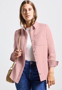 Světle růžová košile s knoflíky a přední kapsou, měkká textura a vyhrnuté rukávy, spojená s bílým topem a modrými džínami.