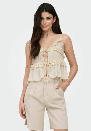 Kobieta z długimi ciemnymi włosami, ubrana w kremowy, falbaniasty top bez rękawów i beżowe szorty z wysokim stanem, pozująca z jedną ręką w kieszeni.