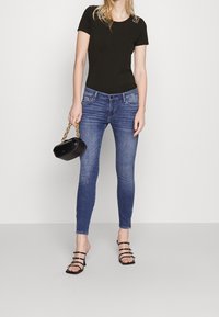 Svart T-shirt med kort ärm och logodetalj, blå slim-fit jeans, svart handväska med kedjehandtag, och svarta sandaletter.