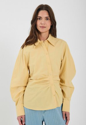 Sárga pamut ing, galléros nyakkal, gombos elülső záródással és deréknál ráncolt részlettel. Hosszú, ballon ujjú és sima textúrájú.