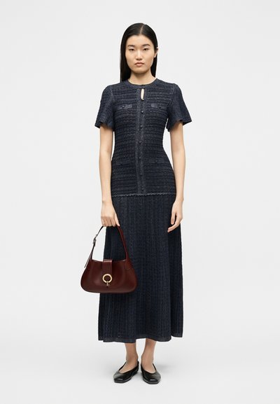 Μια γυναίκα στέκεται φορώντας μπλε ναυτικό κοντομάνικο τοπ με υφή και φούστα που ταιριάζει, κρατώντας μια σκουρόχρωμη κόκκινη τσάντα, με μαύρα φλατ παπούτσια σε λευκό φόντο.