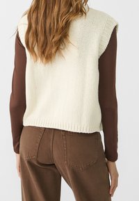 Vrouw met lang haar die een mouwloze crèmekleurige gebreide vest over een bruin shirt met lange mouwen en een bruine broek draagt, gezien van achteren.