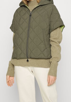 Kvinde iført olivengrøn quiltet ærmelløs hættetrøje over beige langærmet sweater med cremefarvede bukser, stående mod ensfarvet baggrund.