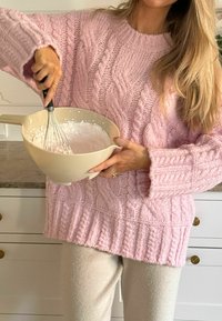 Rozā adīta džinsa ar kabelīšu adījumu, saskaņota ar krēmkrāsas biksēm. Tur rokās bēša maisīšanas trauku, kas piepildīts ar gaisīgu baltu maisījumu.