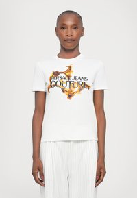 Balta kokvilnas t-kreklu ar krāsainu baroka stila drukājumu, kurā centrā ir izcēlts "VERSACE JEANS COUTURE" biezā melnā burtā.