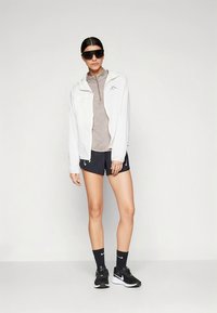 Witte lichtgewicht jas, bruin shirt met kwartrits, zwarte korte broek, zwarte sneakers en sokken. Donkere zonnebril maakt de sportieve outfit compleet.