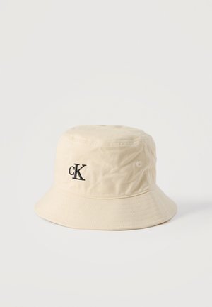 Beige puuvillainen kalastajanhattu, jossa musta "cK"-logo edessä kirjailtuna, tikattu lieriosa ja sivuilla ilma-aukot.