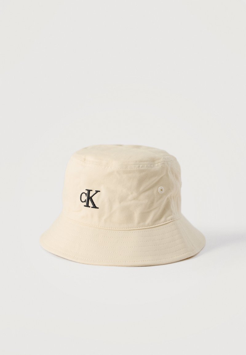 Μπέζ βαμβακερό καπέλο τύπου bucket με μαύρο κέντημα λογότυπου "cK" στο μπροστινό μέρος, με ραμμένη κορυφή και οπές αερισμού στη μία πλευρά.
