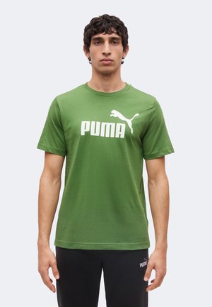 Bărbat stă în picioare, cu fața spre înainte, poartă un tricou verde Puma cu logo alb și pantaloni negri, cu un fundal luminos și simplu.