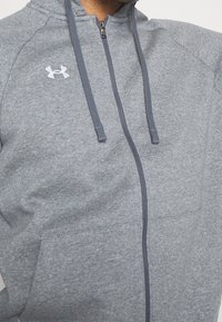 Sudadera gris con cremallera, hecha de tela de felpa suave, presenta un diseño bicolor con cremalleras en gris oscuro y un logo blanco en el pecho.