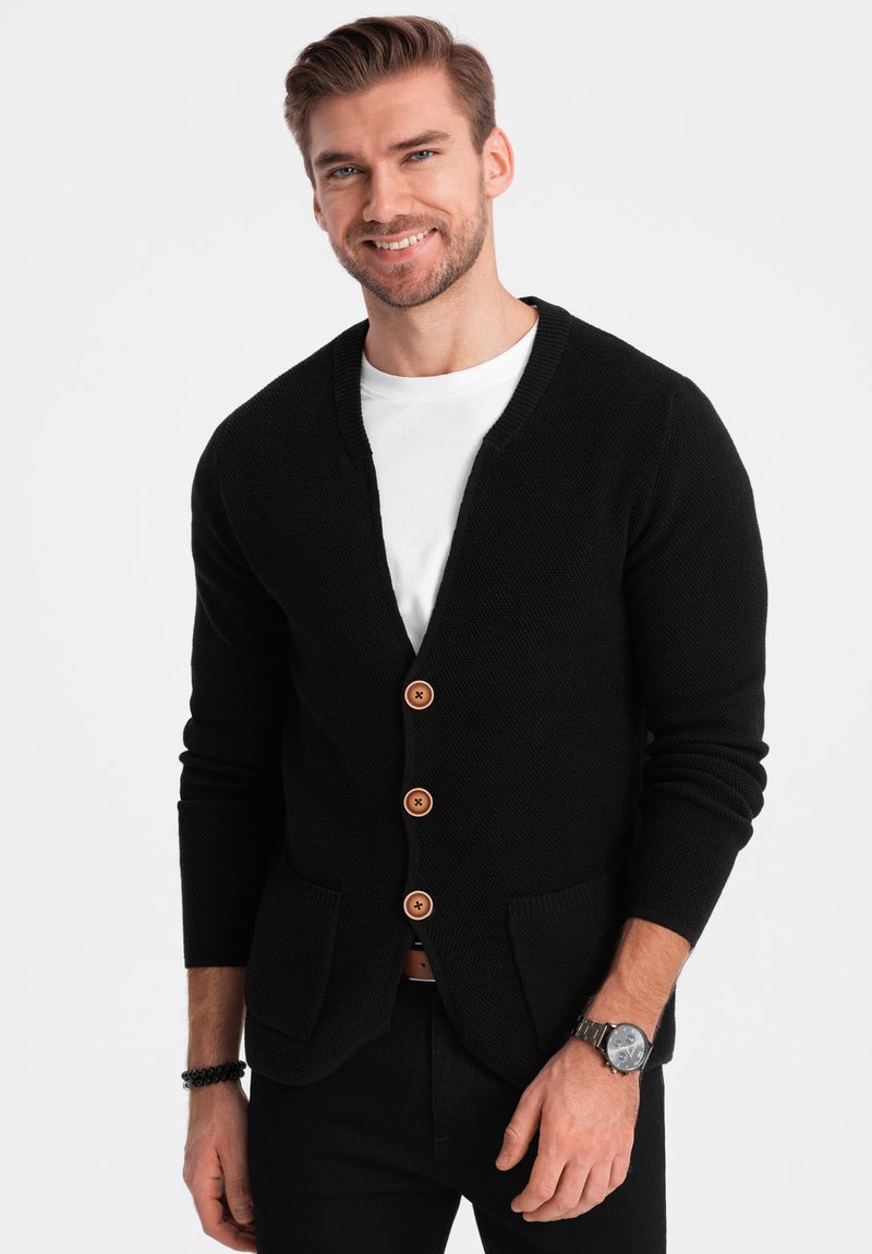 Cárdigan negro de punto con escote en forma de V, que cuenta con cinco botones de madera, dos bolsillos frontales y puños acanalados. Lleva puesto sobre una camiseta blanca.