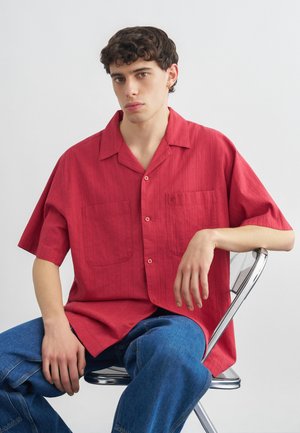 Νεαρός με σγουρά μαλλιά κάθεται σε μεταλλική καρέκλα, φοράει άνετο κόκκινο πουκάμισο με κοντά μανίκια και μπλε τζιν απέναντι σε ένα απλό φόντο.