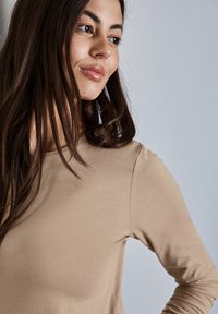 Beige Langarmtop mit rundem Ausschnitt und weicher Textur, das eine glatte Oberfläche und eine taillierte Form aufweist, teilweise von einer Person getragen.