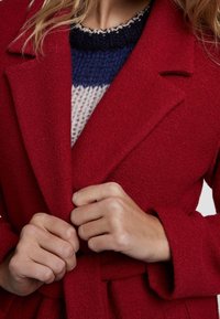 Manteau en laine rouge à la texture douce, avec une taille ceinturée et de larges revers. Des mains tiennent le manteau fermé, laissant entrevoir un pull coloré en dessous.