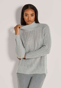 Frau in einem hellgrauen, gerippten Pullover mit Rollkragen und grauen Hosen, die den Kragen des Pullovers mit einer Hand hält.