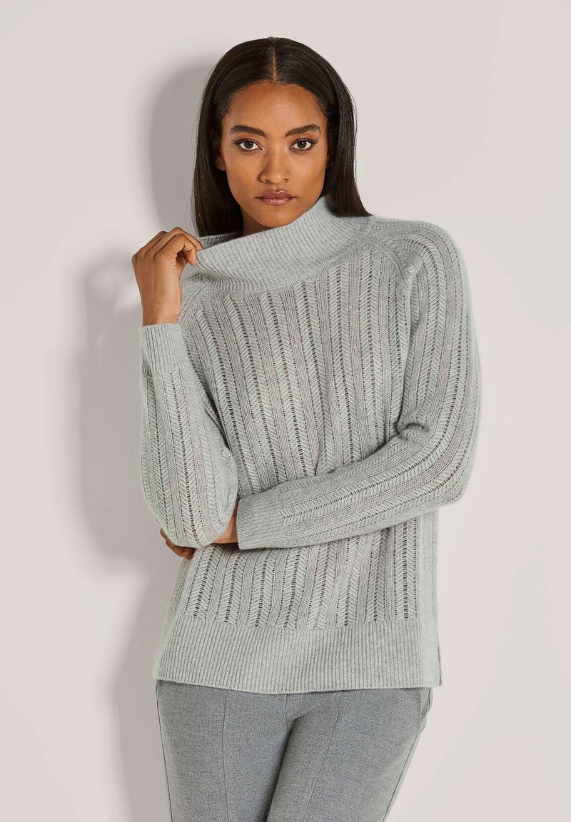 Frau in einem hellgrauen, gerippten Pullover mit Rollkragen und grauen Hosen, die den Kragen des Pullovers mit einer Hand hält.