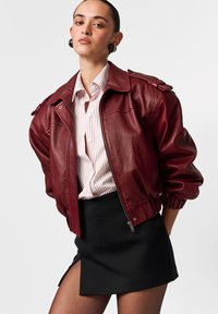 Jachetă bomber din piele vișinie cu închidere cu fermoar, epoleți pe umeri și manșete striate. Asortată cu o cămașă în dungi și o fustă mini neagră.