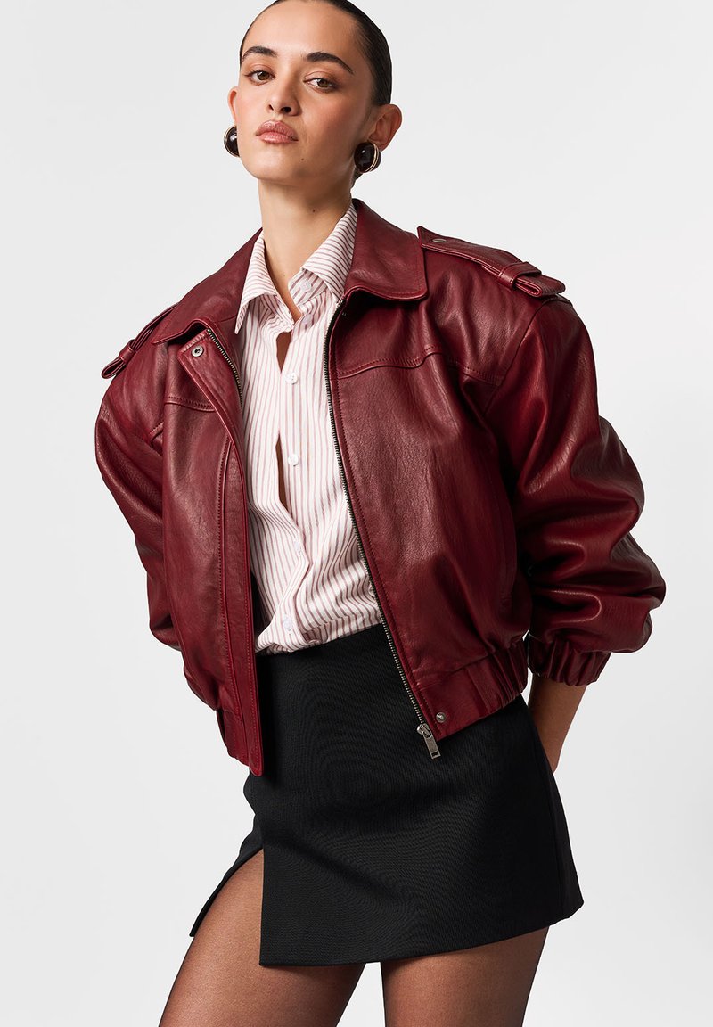 Jachetă bomber din piele vișinie cu închidere cu fermoar, epoleți pe umeri și manșete striate. Asortată cu o cămașă în dungi și o fustă mini neagră.
