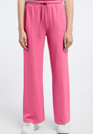 Kobieta w luźnych różowych spodniach dresowych ze ściągaczem i białych tenisówkach, stojąca na tle jednolitego tła.