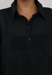 Czarna koszula z kołnierzykiem, trzema guzikami i kieszenią na piersi, noszona przez osobę widoczną od szyi do górnej części klatki piersiowej.