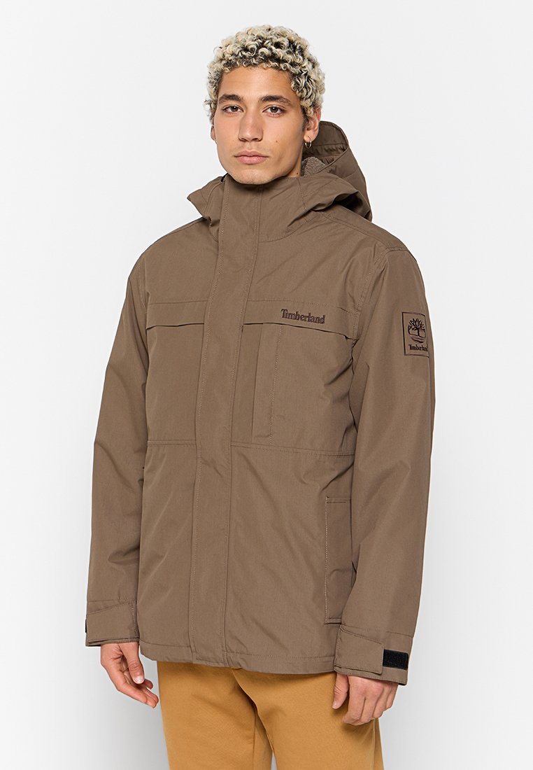 Brązowa wodoodporna kurtka z kapturem, zapinana na zamek błyskawiczny, z dwiema kieszeniami na piersi oraz naszywką z logo Timberland na rękawie. Gładka tekstura.