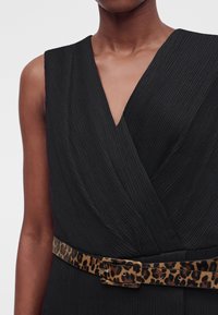 Gros plan d’un torse portant une robe noire texturée sans manches avec une ceinture à motif léopard à la taille.