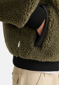 Veste en polaire vert olive avec un extérieur texturé et duveteux, dotée d'un poignet côtelé noir et d'une doublure intérieure visible à la poche ouverte.