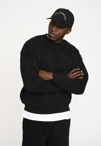 Sort sweatshirt med rund hals og ribbede manchetter, med et diskret logo. Sammen med en hvid skjorte og en sort baseballkasket.