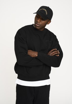 Sort sweatshirt med rund hals og ribbede manchetter, med et diskret logo. Sammen med en hvid skjorte og en sort baseballkasket.