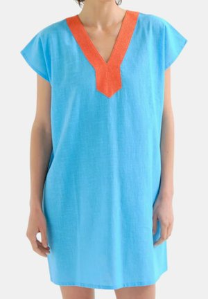 Donna che indossa un vestito blu largo a maniche corte con una profonda scollatura a V bordata di arancione brillante, in piedi su uno sfondo bianco.
