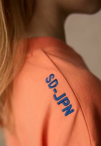 Orange stickad sweatshirt med rund hals, försedda med blå broderad text "SD_JPN" på vänster axel. Mjuk textur, figurnära passform.