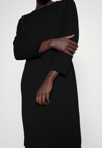 Černé dlouhé šaty s volným střihem, zdobené texturovanými akcenty na ramenou a lodičkovým výstřihem, vyrobené z hladkého materiálu.