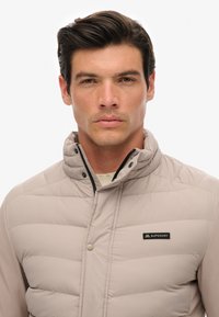 Veste rembourrée beige clair avec un col montant, fermeture zippée, et un détail de logo sur la poitrine. Comprend des sections cousues pour un effet de texture supplémentaire.
