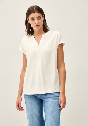 Blusa bianca a maniche corte con scollo a V e vestibilità rilassata. Realizzata in tessuto morbido, presenta cuciture minime all'orlo. Abbinata a jeans azzurri chiari.