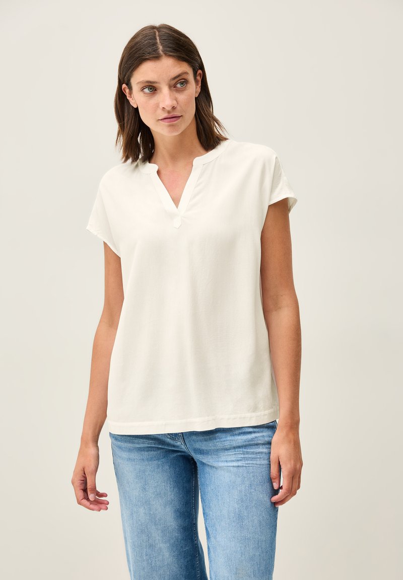 Blouse blanche à manches courtes avec un col en V et une coupe décontractée. Fabriquée en tissu doux, elle présente des coutures minimales à l'ourlet. Associée à un jean bleu clair.