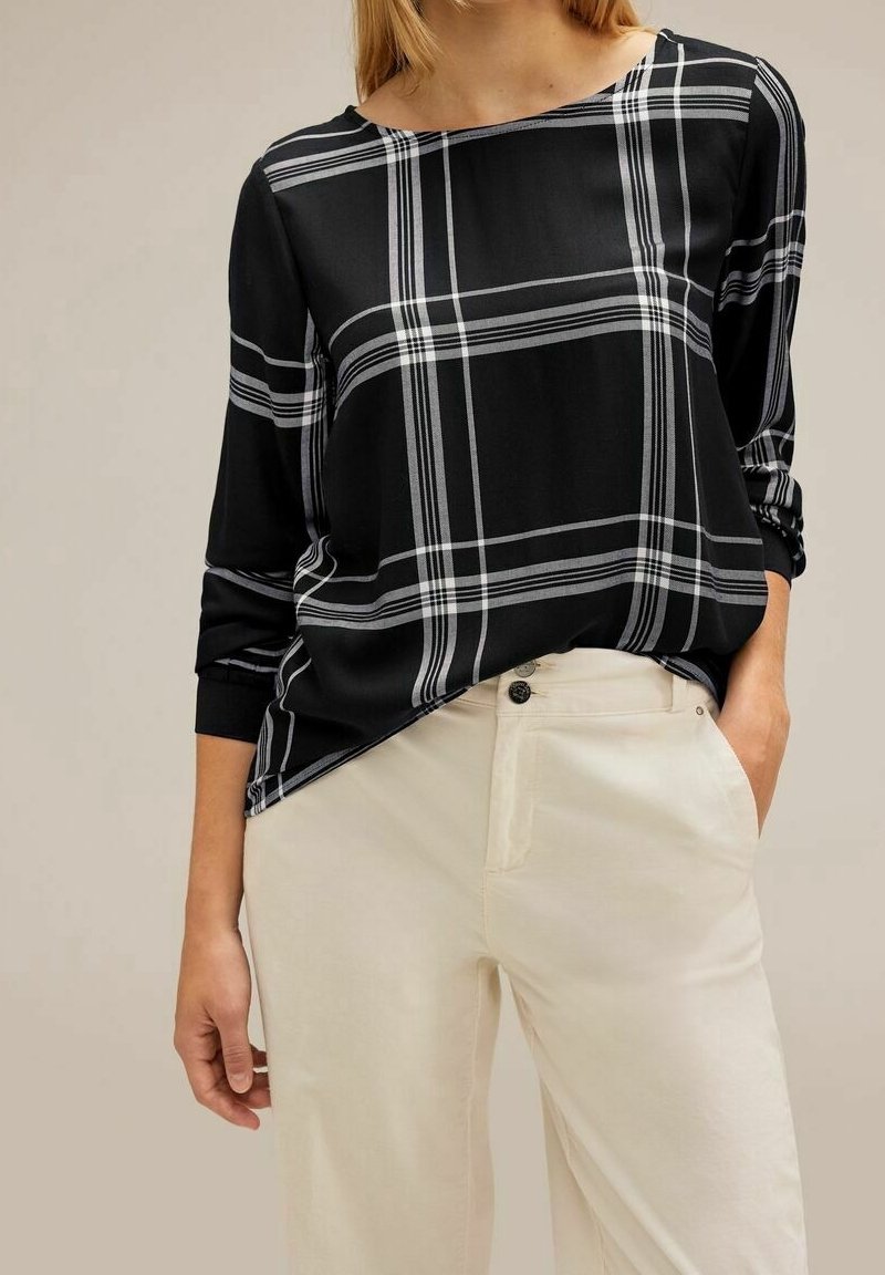 Blusa a scacchi in bianco e nero con maniche lunghe, caratterizzata da una vestibilità ampia e scollo a barca. Abbinata a pantaloni color crema, con dettagli in oro visibili sui bottoni.
